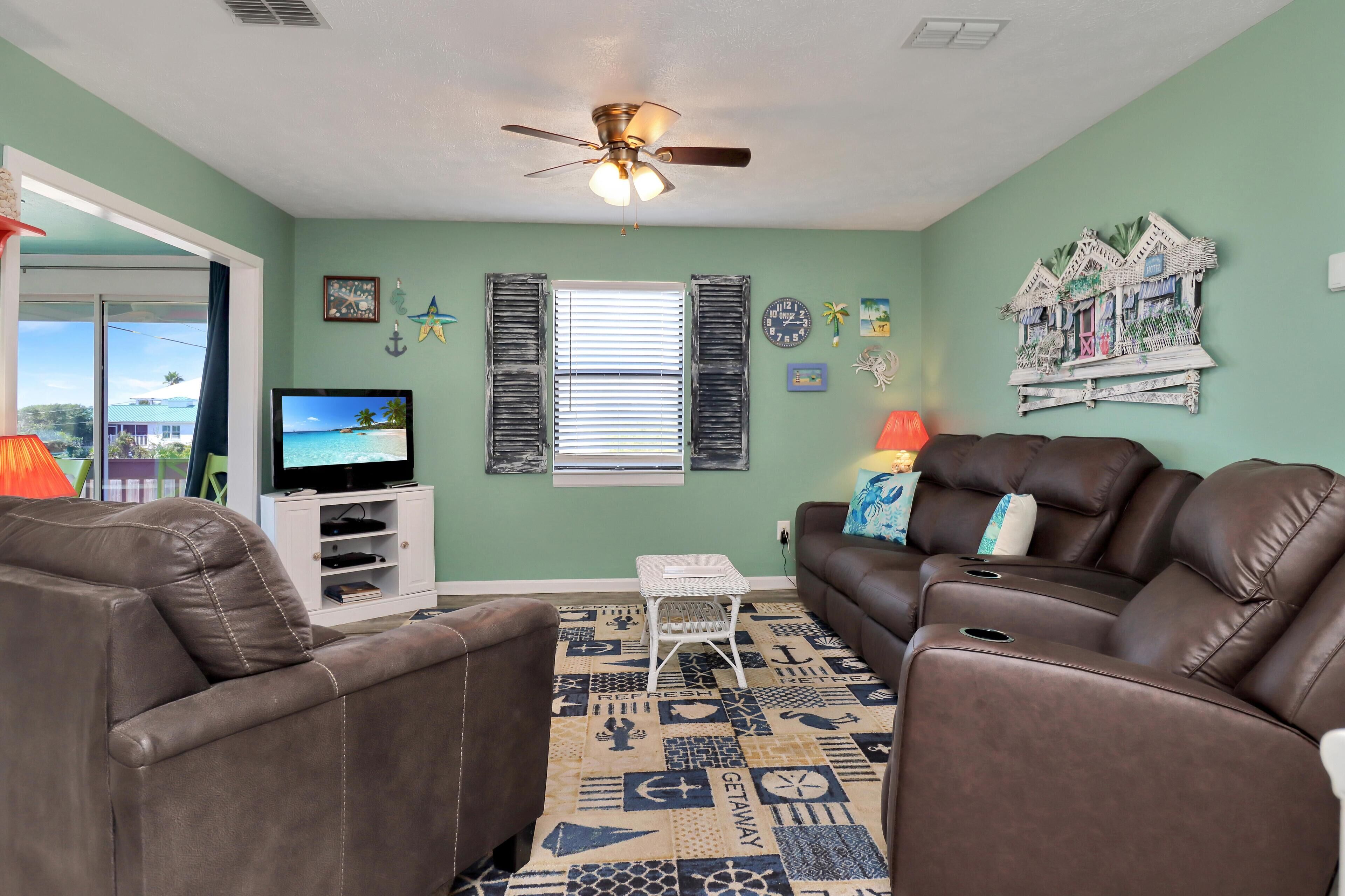 A living room with a brown leather sofa, a white coffee table, and a television on a white stand. There is a ceiling fan, a window with blinds, and a decorative wall piece.
