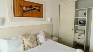 Ein ordentlich bezogenes Bett mit weißen Bettwäsche, ein Deckenventilator, ein hölzernes Kunstwerk darüber, ein Nachttisch mit einer Pflanze und ein Kleiderschrank mit einem Radio.