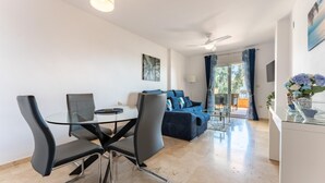 A modern living room with a glass dining table, black chairs, a blue sofa, and a ceiling fan. - CT 232 - Eden Golf Sur - Southfacing Apartment (Mijas)