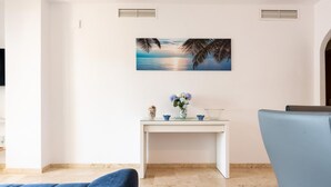 A modern living room with a white wall, a painting of a beach scene, a small white table with decorative items, and a blue sofa. - CT 232 - Eden Golf Sur - Southfacing Apartment (Mijas)