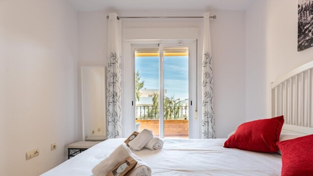 A hotel room with a bed, two red pillows, a white curtain, and a view of a balcony with a railing. - CT 232 - Eden Golf Sur - Southfacing Apartment (Mijas)