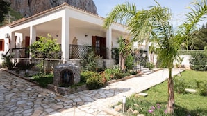 A white building with a tiled roof, a stone pathway, and a garden with palm trees and shrubs.