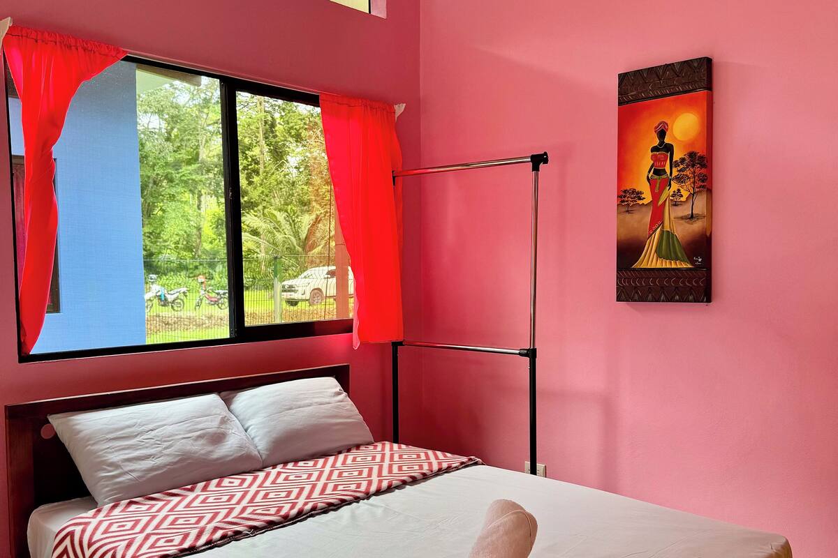 A bedroom with a bed, a window with red curtains, a painting on the wall, and a view of greenery outside.