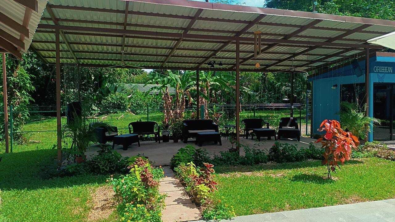 Un área de estar al aire libre cubierta con mesas y sillas, rodeada de follaje y un pequeño edificio con la leyenda 'CARIBBEAN'.