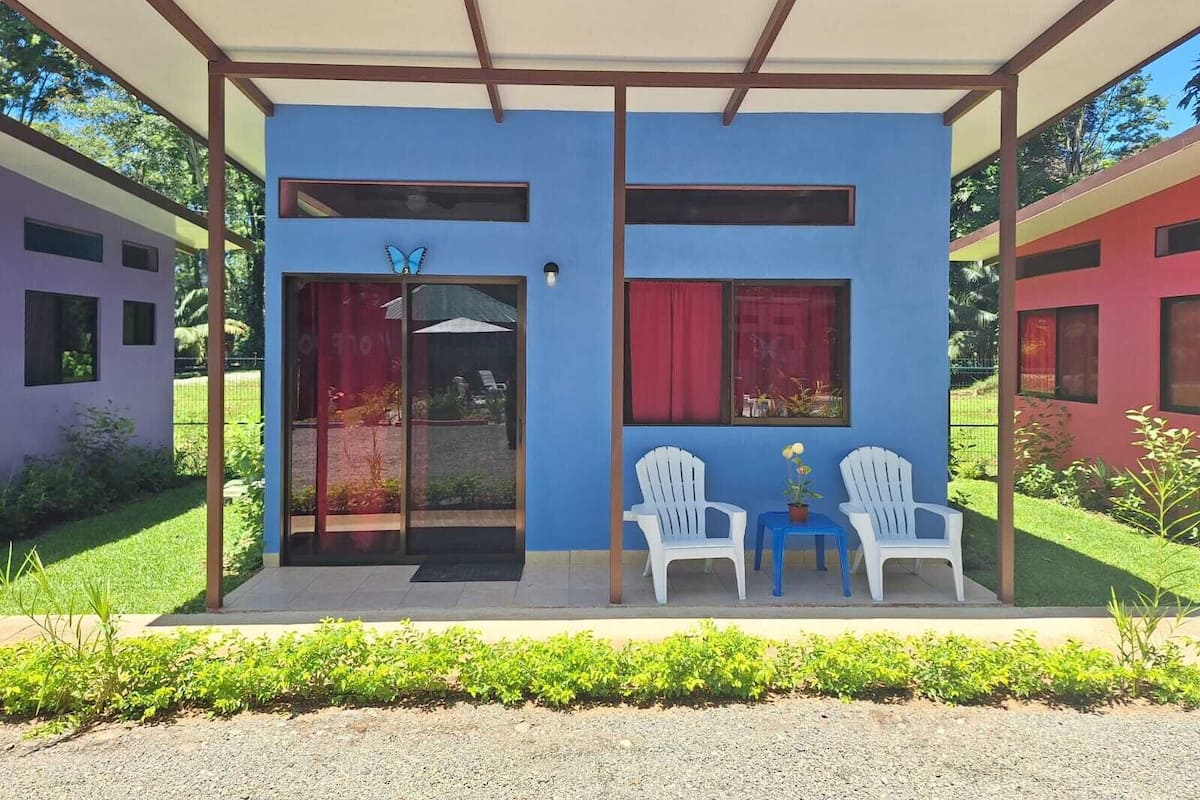 A modern building with a blue facade, two white chairs under a covered patio, and a small table with a potted plant.