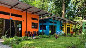 Een modern gebouw met een levendige oranje gevel, een blauw gedeelte en een geel gedeelte. Er zijn terrassen met stoelen en tafels, en een verzorgde tuin met diverse planten en bloemen.