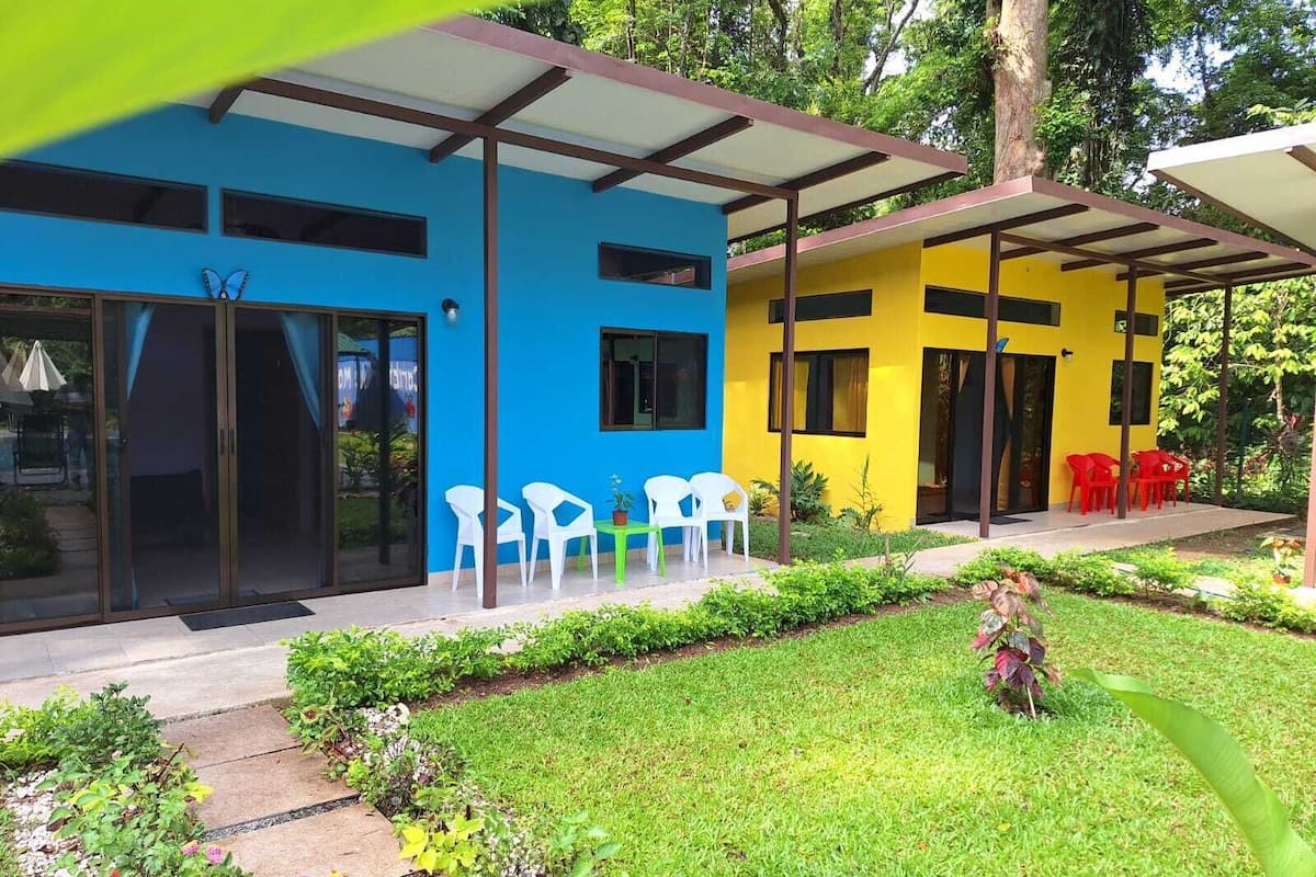 A modern building with a blue and yellow facade, large glass doors, and a covered patio area with white and red plastic chairs.