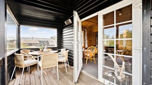 Une terrasse en bois avec des chaises et une table en osier, une porte vitrée donnant sur un espace intérieur, et une vue sur une maison en arrière-plan.