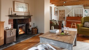 A cozy living room with a fireplace, wooden flooring, a green armchair, and a coffee table with books and glasses. - Calenick -  a stylish cottage that sleeps 6 guests  in 3 bedrooms (Trelights)