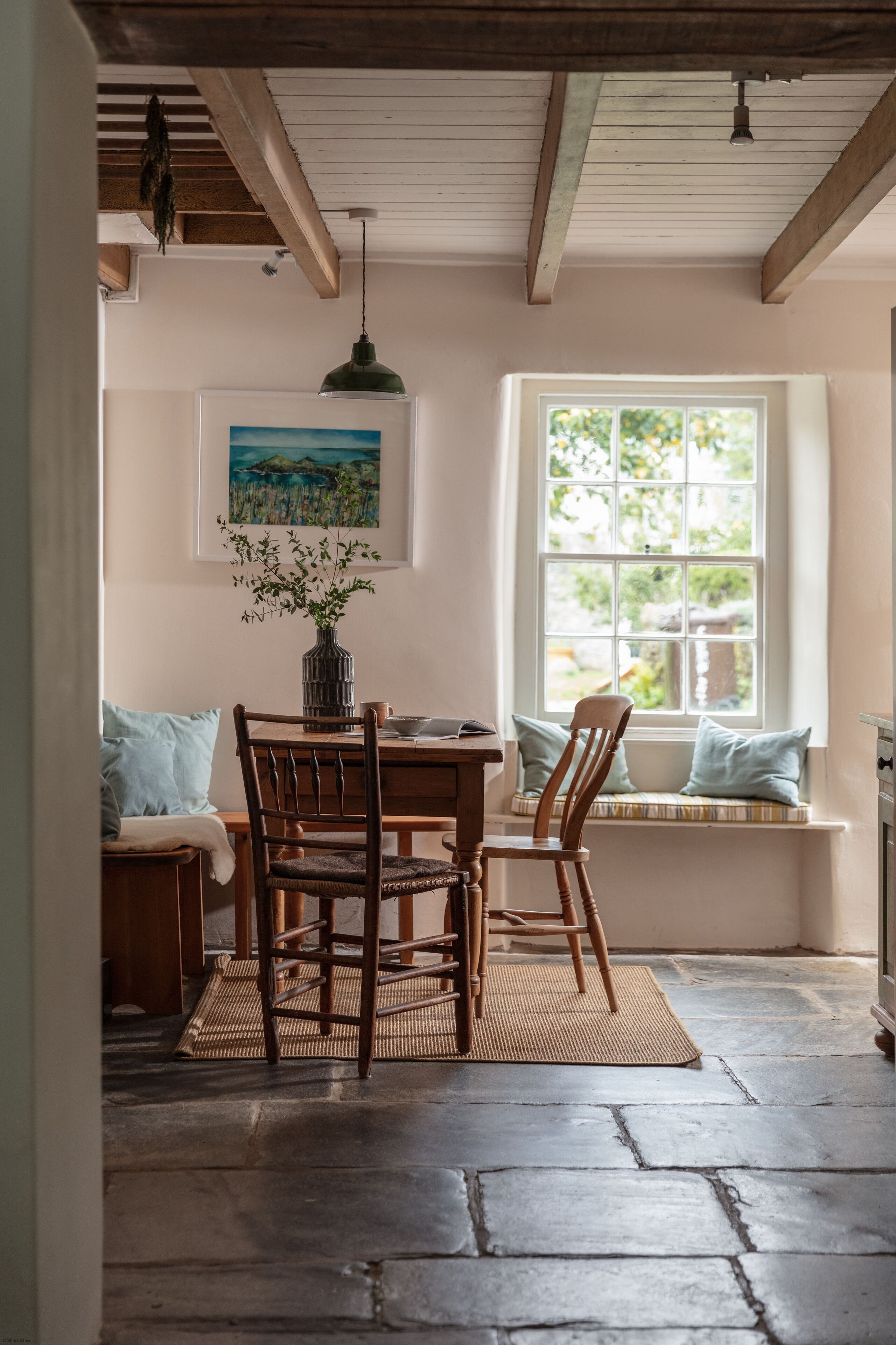 A dining area with a wooden table, chairs, a woven mat, a hanging pendant light, a painting, and a window with a view of greenery.