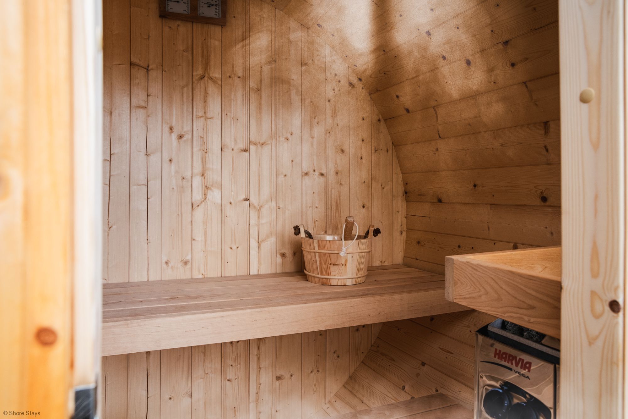 A wooden sauna room with a wooden bench, a bucket with items, and a Harvia VSGA sauna head.