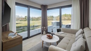 Ein modernes Wohnzimmer mit einer Couch, einem Couchtisch und einem Fernseher. Große Fenster bieten Blick auf eine Rasenfläche und Bäume.