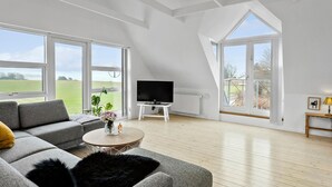 Ein geräumiges Wohnzimmer mit großem Fernseher, grauem Sofa und Blick auf ein grünes Feld durch große Fenster.