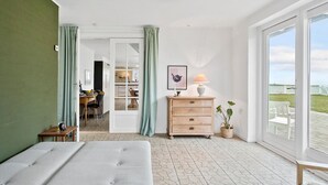 Ein Zimmer mit einem Bett, einem Holzschrank, einem Schreibtisch mit Stühlen, einem Esstisch und Blick ins Freie durch ein großes Fenster.