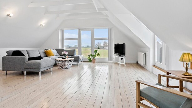 Ein großzügiger Loftsraum mit hoher Decke, großen Fenstern mit Blick auf die Landschaft, einem grauen Sofa, einem Holztisch und einem Fernseher.