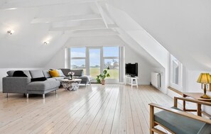 Een ruime loft met een hoge plafond, grote ramen met uitzicht op het platteland, een grijze bank, een houten salontafel en een televisie.