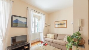 A living room with a sofa, a glass coffee table, a TV on a stand, a plant, and a window with curtains.