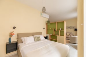 A bedroom with a bed, bedside tables, a hanging light fixture, a green shelving unit, and a sofa in the adjacent living area.