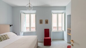 A bedroom with a bed, two windows, a red chair, and a hanging light fixture. - iFlat | Lovely in Trevi Fons Trevi II (Roma)