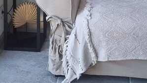 Een hoek van een kamer met een bank, een decoratieve plaid, een decoratieve plank met een buste, en een tafeltje met een ventilator.