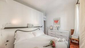 Ein ordentlich bezogenes Bett mit weißen Leinen, ein Nachttisch mit einer Lampe, ein gerahmtes Kunstwerk an der Wand und ein kleiner Holzsessel.