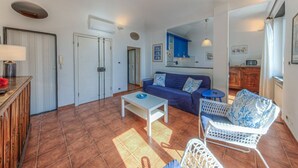 Ein Wohnzimmer mit einer Couch, einem Couchtisch und einem Sessel. Im Hintergrund ist eine Küchenzeile erkennbar.