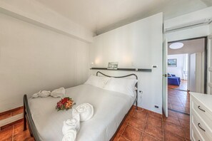 A neatly made bed with white linens and towels, a bedside table with a lamp, and a view into another room with a blue sofa.