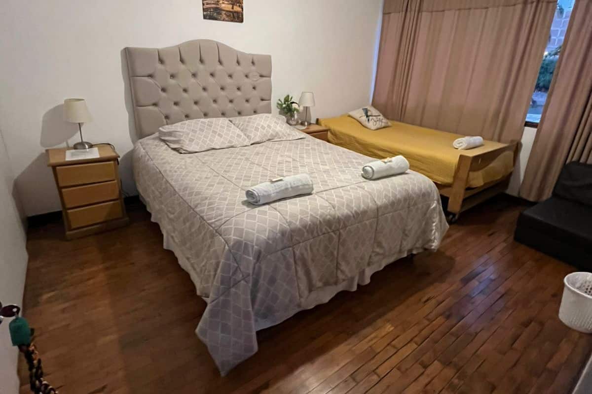Un dormitorio con una cama grande, una mesita de noche con lámpara, una pequeña mesa con una planta y una ventana con cortinas.