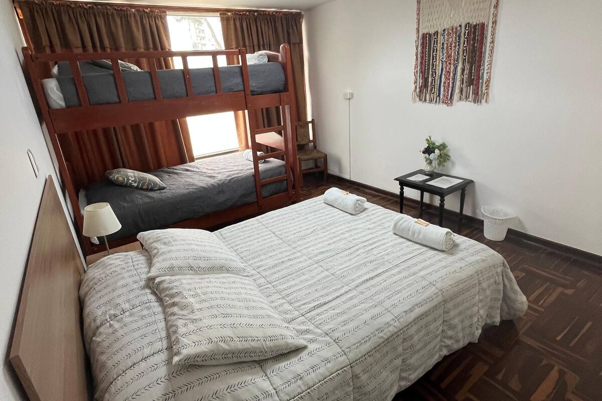Una habitación con literas, un escritorio, una silla, una mesita con una planta y un cuadro en la pared.