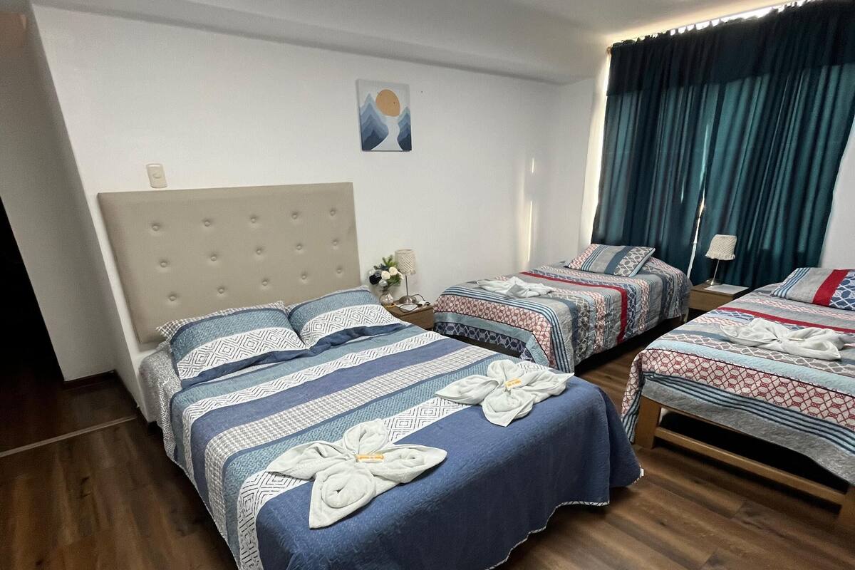 Habitación de hotel con una cama grande, dos camas más pequeñas, suelo de madera, una lámpara de techo, una lámpara de pared, un cuadro y una ventana con cortinas.
