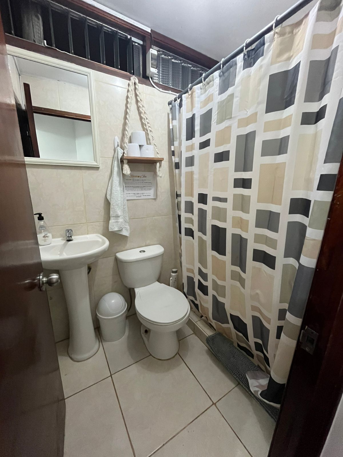 A compact bathroom with a pedestal sink, two toilets, a small trash bin, and a shower curtain with a geometric pattern.