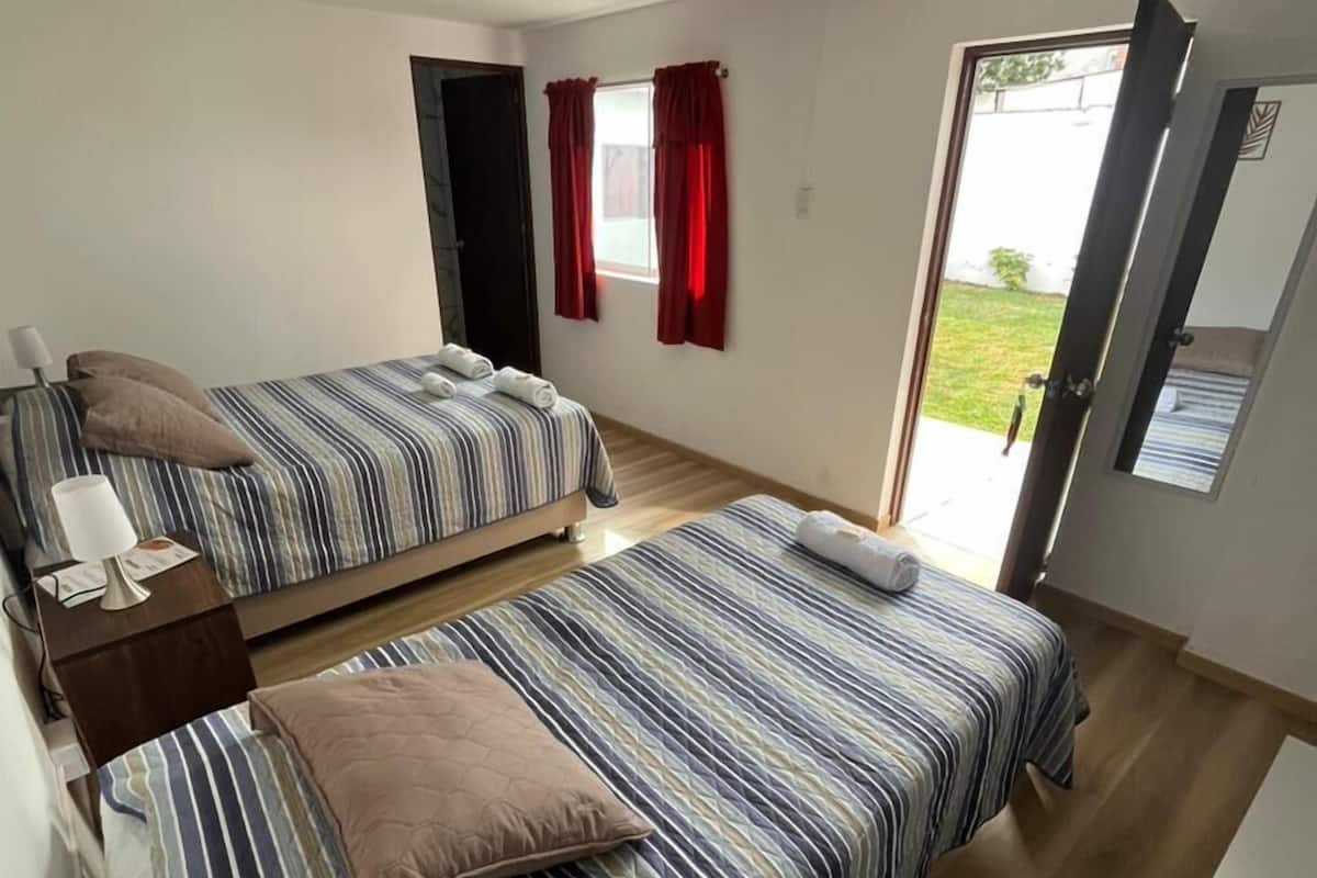 Habitación de hotel con dos camas, una mesita con una lámpara, una ventana con cortinas rojas y una puerta que da al exterior.