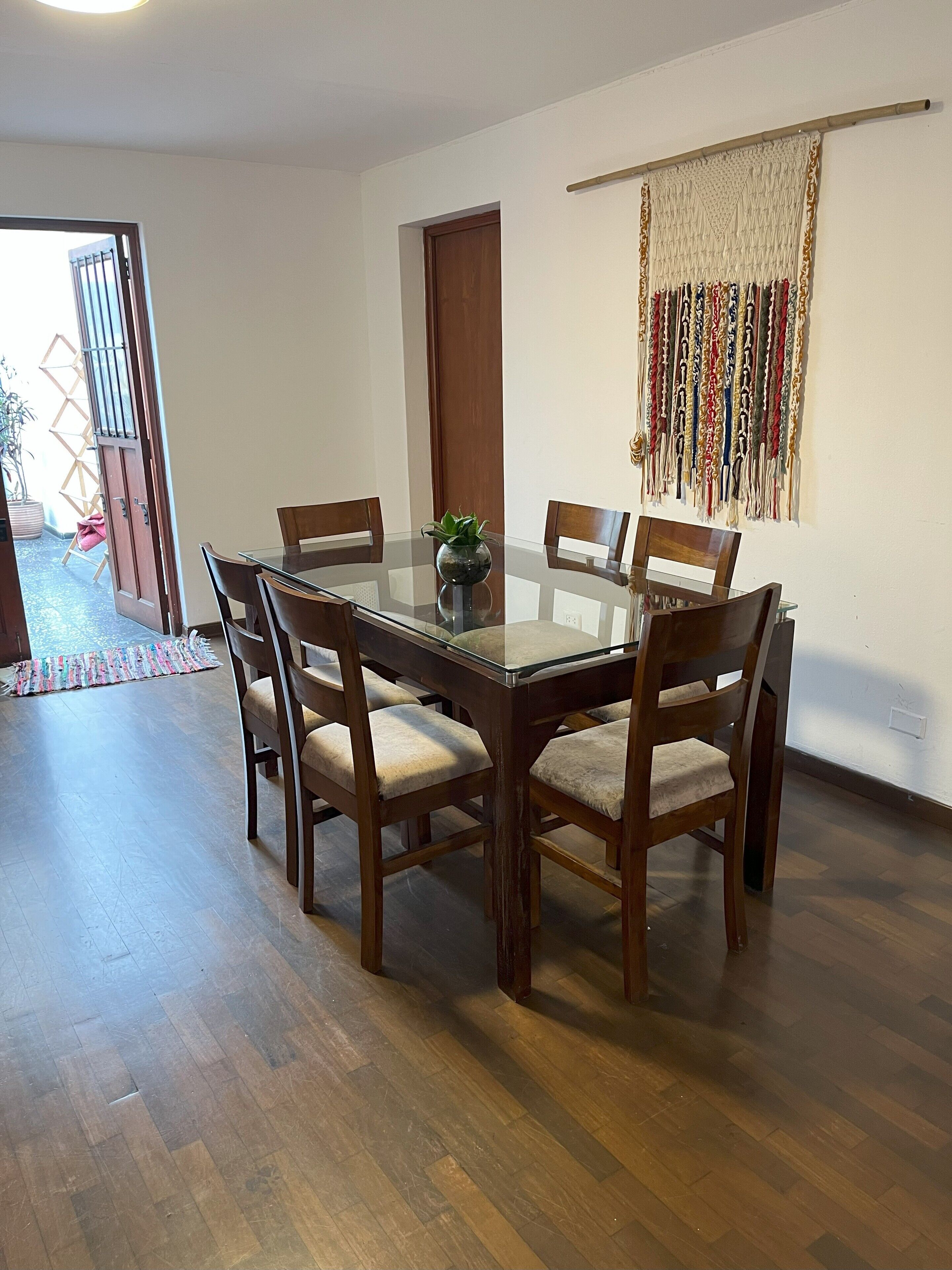 A dining area with a glass-top table surrounded by wooden chairs, a potted plant, and a decorative wall hanging.