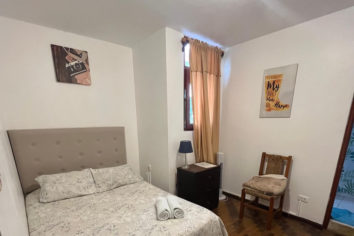Un dormitorio con una cama, una silla, una mesita de noche, una lámpara, una ventana con cortinas y un cartel en la pared.
