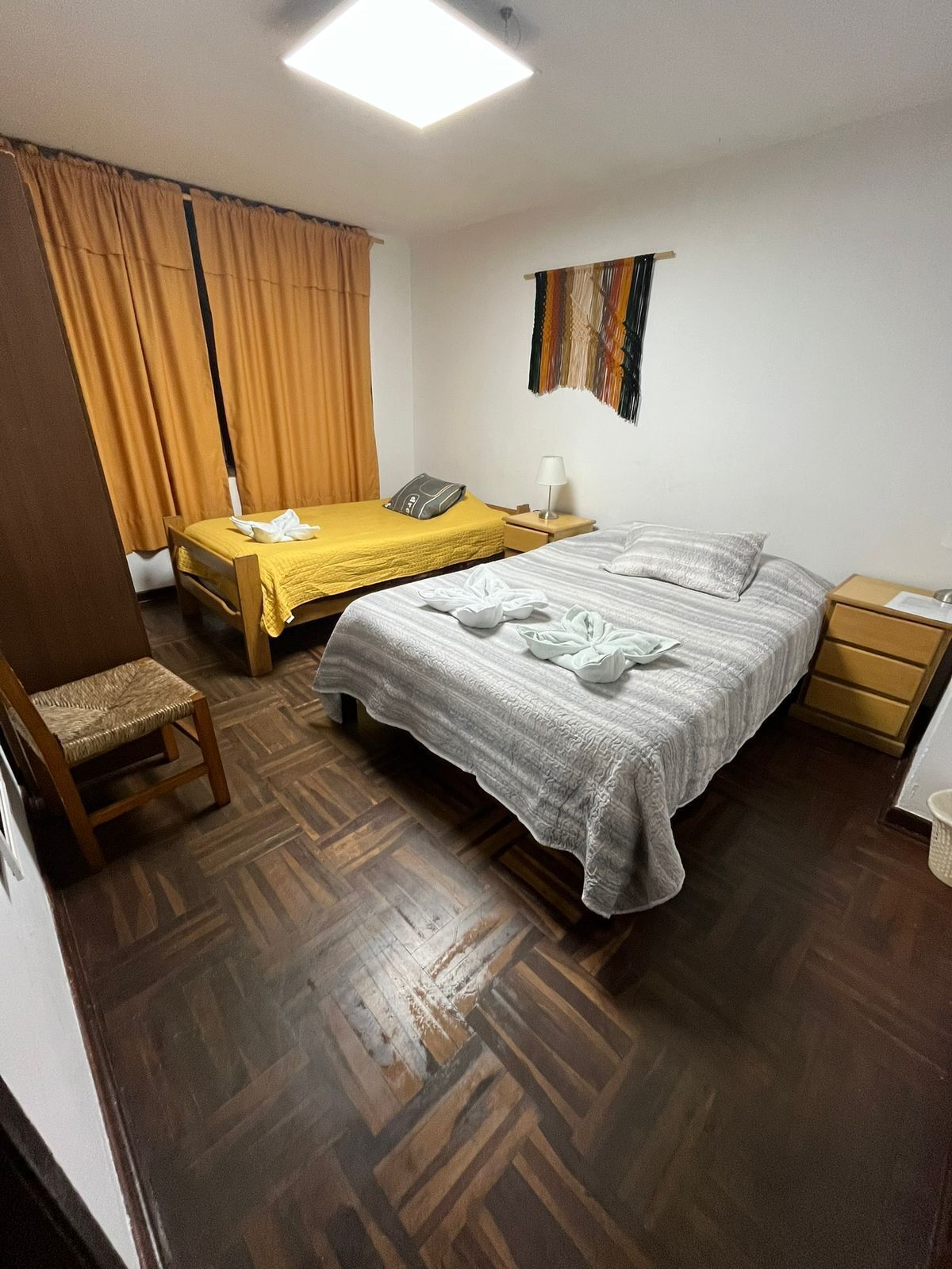 Habitación de hotel con dos camas, una silla de madera, una mesita de noche con lámpara y un adorno en la pared.