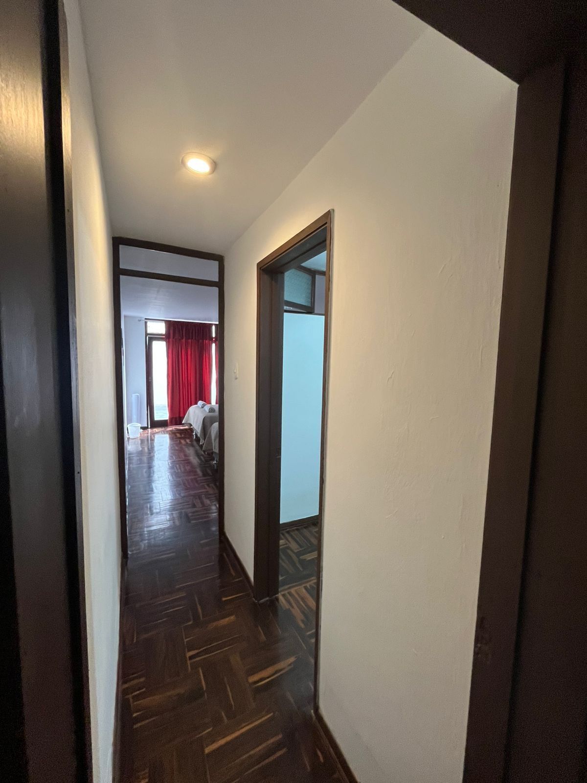 A narrow hallway with a wooden floor leading to a room with a bed and red curtains.