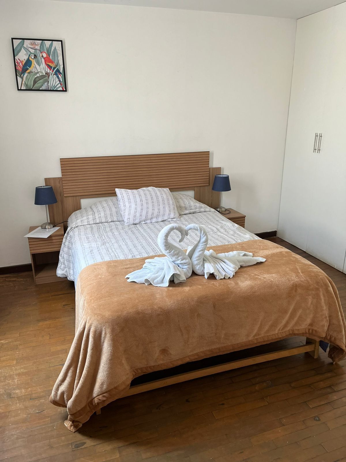 A neatly made bed with a brown blanket and two swan-shaped towels, a wooden headboard, two bedside lamps, and a wall-mounted artwork.