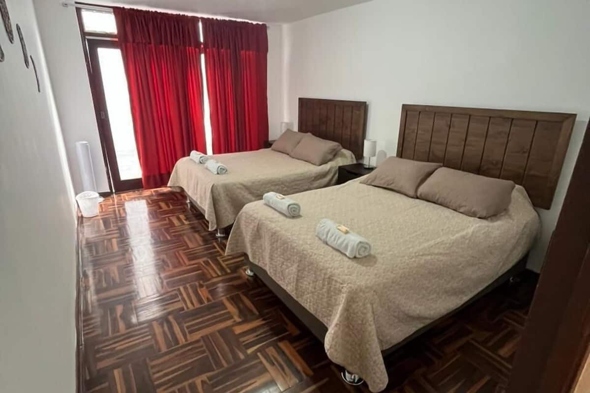 Habitación de hotel con dos camas, suelo de parqué de madera, cortina roja y ventilador de techo.