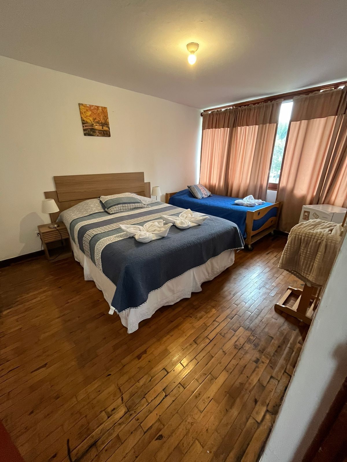 A bedroom with a large bed, a smaller bed, a wooden chair, a nightstand, a lamp, and a painting on the wall.