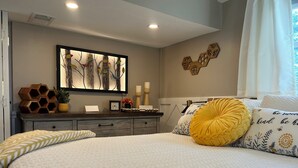 Une chambre à coucher comprenant un lit, des tables de chevet, une commode, un tableau décoratif au mur et une fenêtre avec des rideaux.