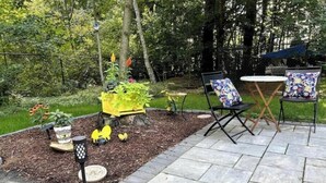 Une terrasse extérieure avec un sol en pierre, deux chaises dotées de coussins à motifs floraux, une petite table et un abreuvoir à oiseaux agrémenté de plantes.