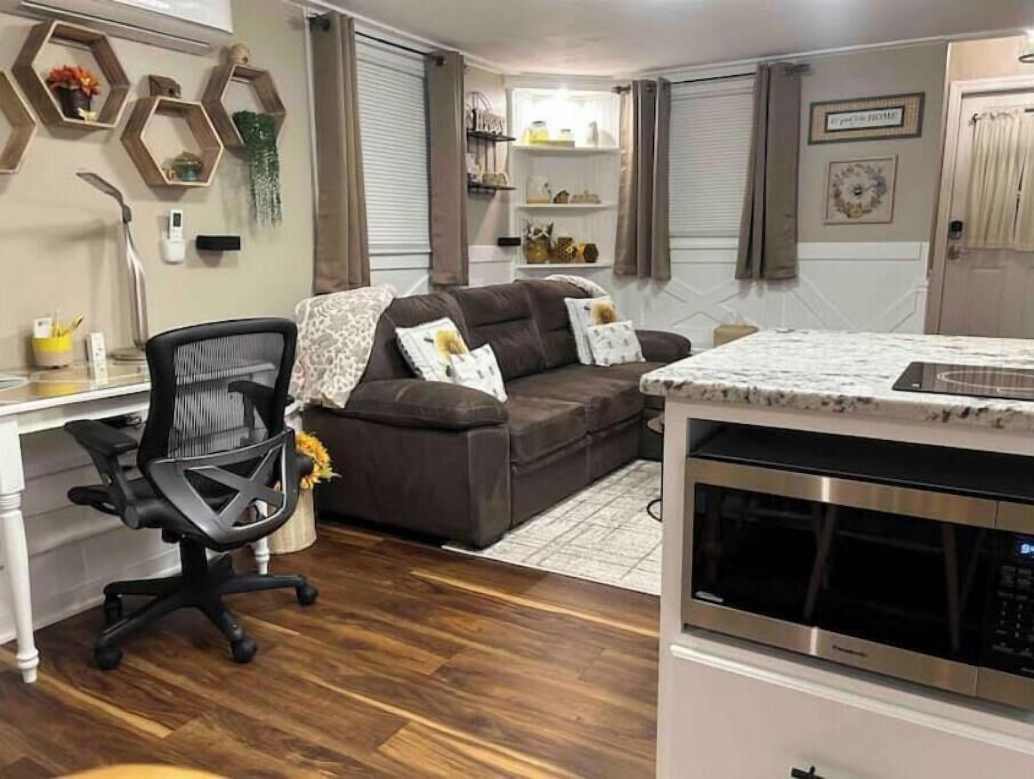 Un espace de vie compact comprenant un coin cuisine, un canapé marron, un fauteuil de bureau noir et des étagères décoratives à motifs hexagonaux.