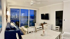 Una sala de estar moderna con un sofá, una mesa de comedor y sillas. Hay un televisor montado en la pared y un balcón con vistas.