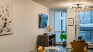 Una sala de estar moderna con una mesa redonda, un sofá, un televisor montado en la pared y un ventanal con vistas a edificios.