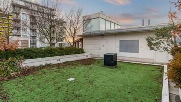 A modern building with a flat roof, a small garden area with grass and a black trash bin, and a clear sky.