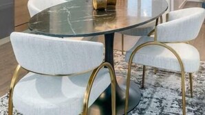 Un comedor moderno con una mesa redonda de cristal, sillas blancas tapizadas y una lámpara de araña dorada.