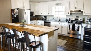 Cocina con alacenas blancas, una isla de madera con taburetes de bar, electrodomésticos de acero inoxidable y una ventana con persianas.