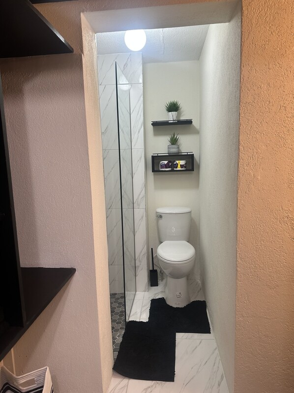 A bathroom with a toilet, a shower, and a small shelf with plants. - West View 3- Modern Studio Apartment (Bridgetown)