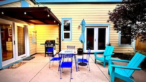 Une terrasse avec des chaises bleues et une table, un barbecue et un espace couvert avec une porte.