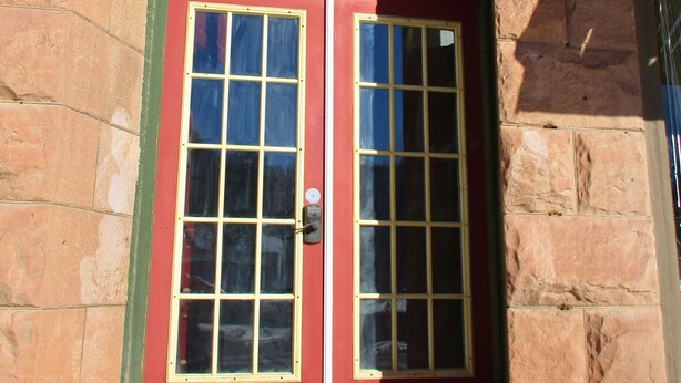 Una puerta doble roja con paneles de vidrio, ubicada en un edificio de piedra con un pequeño escalón que lleva hacia ella.